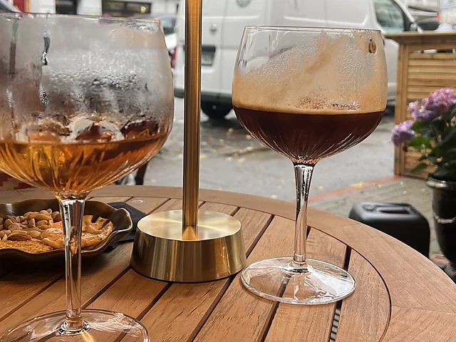Ein Glas Aperitivo mit Eis und ein Glas dunkles Bier auf einem runden Holztisch mit goldener Tischlampe, dahinter die St