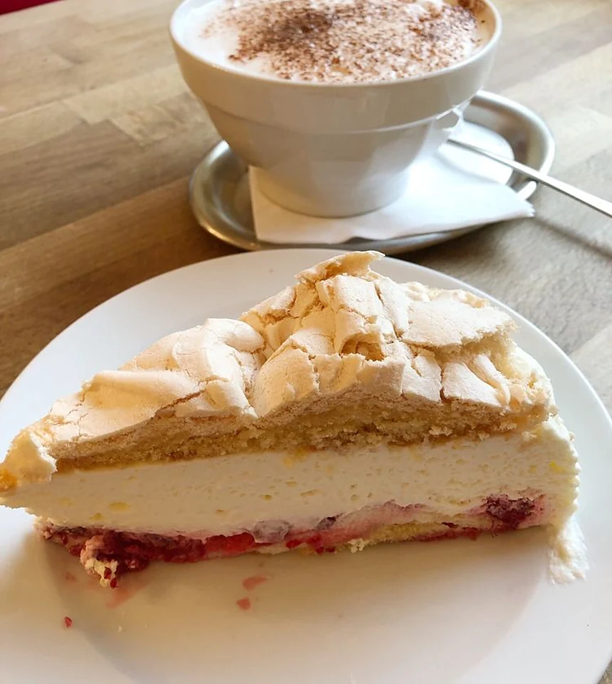 Stück Baiser-Torte mit Fruchtboden auf weißem Teller, daneben ein Cappuccino