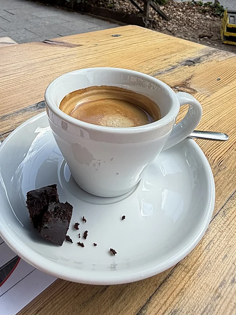 Espresso in einer weißen Tasse mit einem Stück Brownie auf der Untertasse auf einem Holztisch draußen