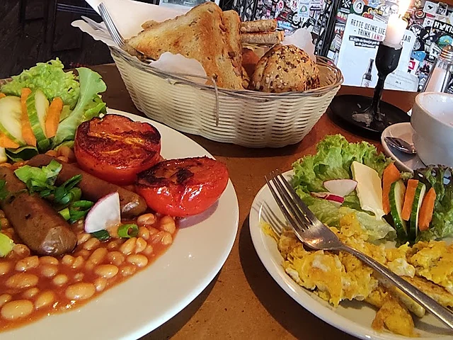 Englisches Frühstück mit Baked Beans, gegrillter Tomate, Würstchen und Salat sowie ein Teller mit Rührei und Gemüsestick