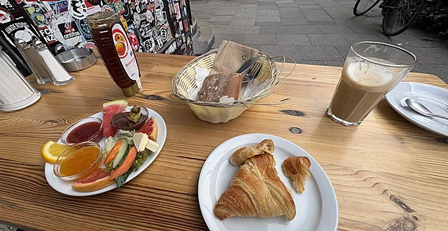 Frühstücksteller mit Obst, Käse und Gemüse, ein Teller mit Croissant, Brotkorb und Latte Macchiato auf einem Holztisch v