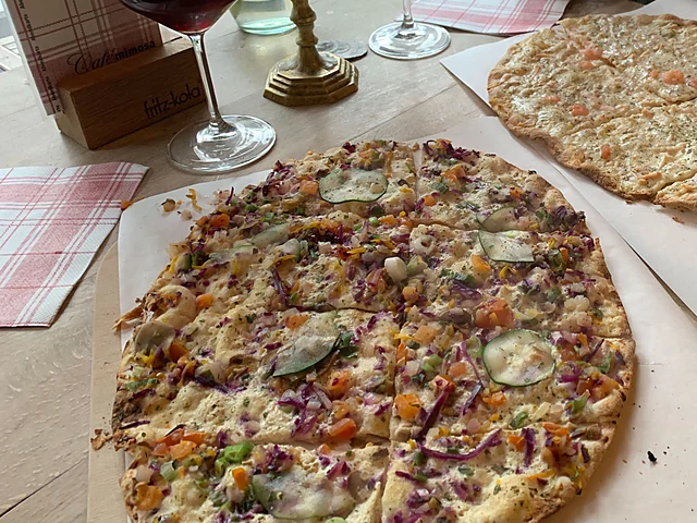 Flammkuchen mit Gemüse, roten Zwiebeln und Käse auf Backpapier, daneben ein zweiter Flammkuchen und Rotweingläser auf karierter Tischdecke