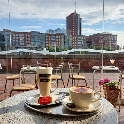 Latte macchiato und Cappuccino auf einem runden Terrassentisch, dahinter der Binnenhafen und Backsteinhäuser