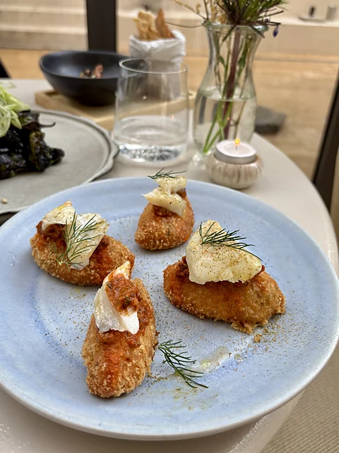 Vier panierte Kroketten mit Dill und Crème auf einem hellblauen Teller, dahinter Kerze, Wasserglas und Blumenvase