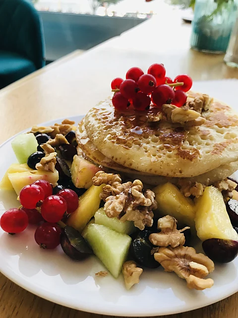 Zwei Pancakes mit roten Johannisbeeren, Walnüssen, Melone, Ananas und Beeren auf weißem Teller am Fenstertisch