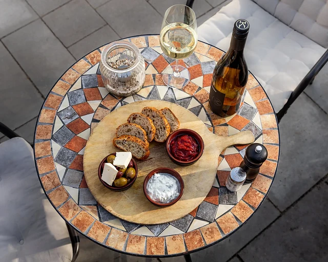Holzbrett mit Brotscheiben, Oliven, Feta, Ajvar und Tsatsiki auf einem Mosaiktisch, daneben ein Glas Weißwein und eine Flasche