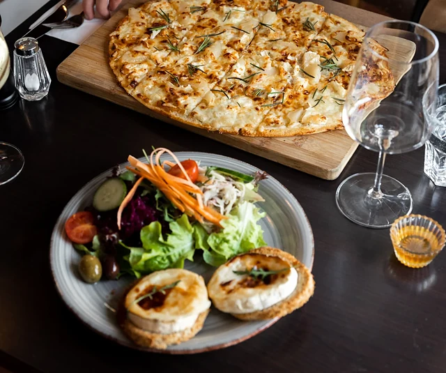 Flammkuchen mit Ziegenkäse und Rosmarin auf einem Holzbrett, davor ein Teller mit Salat und gratiniertem Ziegenkäse