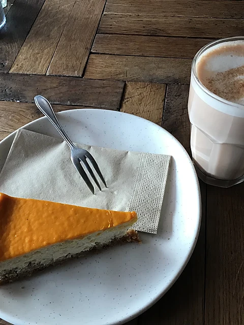 Ein Stück Kürbis-Cheesecake mit Gabel auf einem weißen Teller, dahinter ein Latte im Glas auf Parkettboden-Tisch