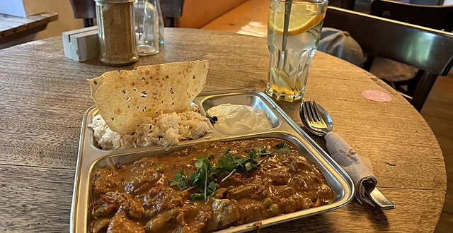 Curry mit Reis, Joghurt und Papadam auf einem Edelstahltablett auf einem Holztisch, dahinter ein Glas Wasser mit Zitrone