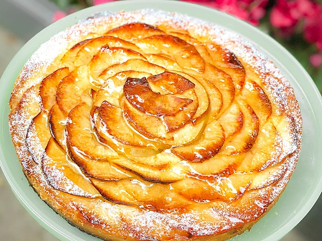 Apfeltarte mit rosettenförmig gelegten Apfelscheiben und Puderzucker auf grünem Teller, dahinter rosa Blüten