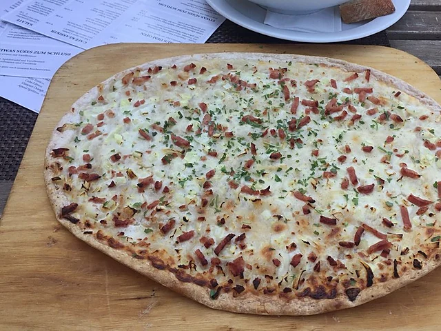 Flammkuchen mit Speck und Zwiebeln auf einem Holzbrett auf einem Holztisch, dahinter eine Schale Salat