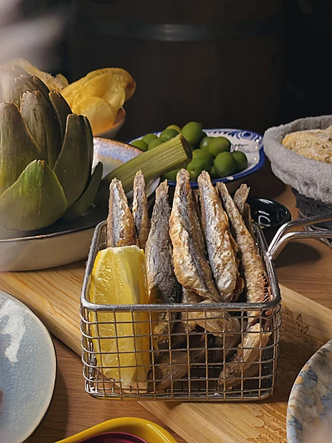 Frittierte Sardinen mit Zitrone in einem Drahtkörbchen, dahinter Artischocke, Oliven und Brotkorb