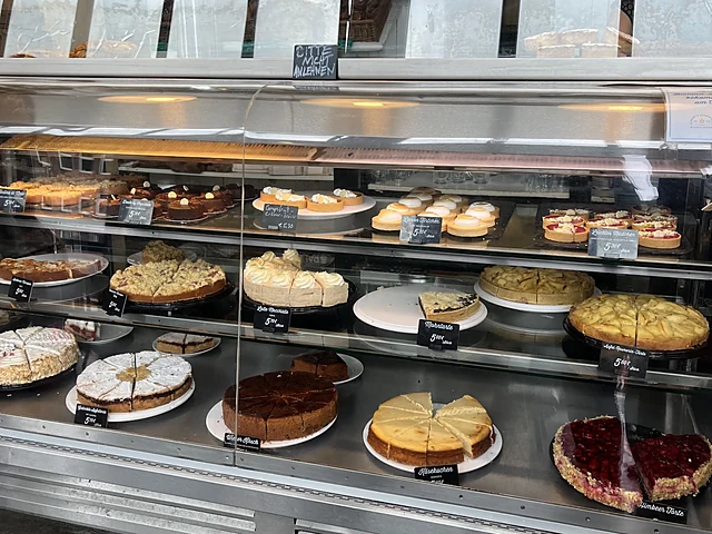 Kuchentheke mit angeschnittenen Torten, Käsekuchen, Himbeertorte und Streuselkuchen hinter Glas, darüber Regale mit Brot