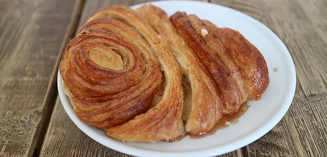Ein Franzbrötchen mit karamellisierter Zimtkruste auf weißem Teller auf einem Holztisch