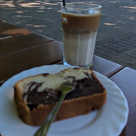 Latte macchiato im Glas und ein Stück Marmorkuchen mit Gabel auf weißem Teller auf der Terrasse, dahinter der Park