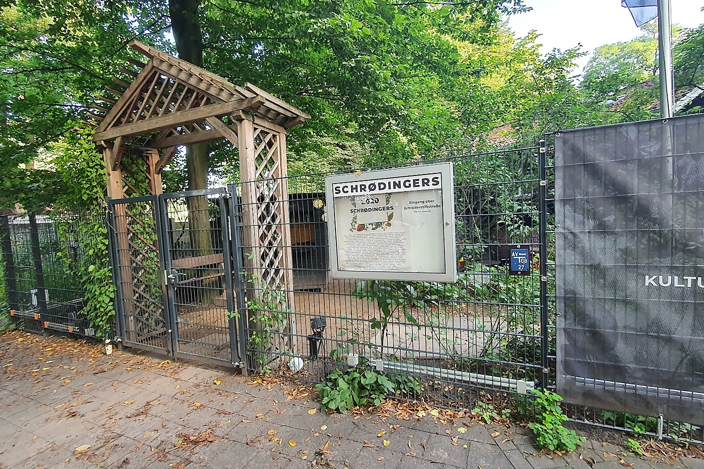 Eingangstor mit Holzpergola, dahinter das Schild Schrødingers und ein Banner mit der Aufschrift Kulturzentrum, ringsum G