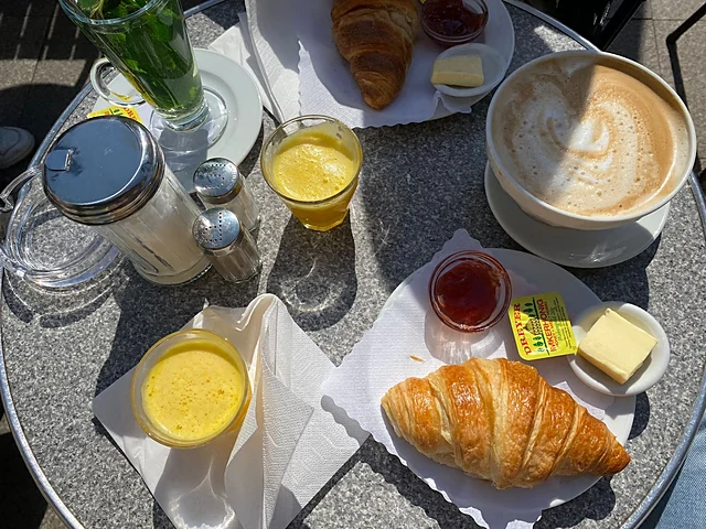 Frühstück von oben auf einem Marmortisch: Croissant, Butter, Konfitüre, Orangensaft, Cappuccino und Eierlikörgläser in der Sonne