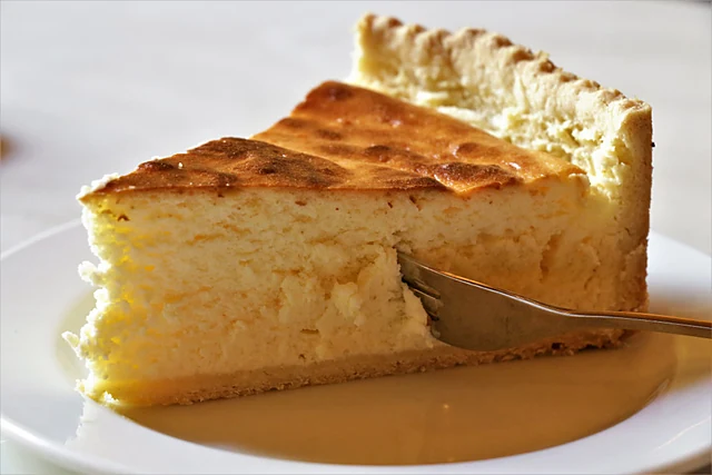 Ein Stück Käsekuchen auf weißem Teller mit Gabel, im Hintergrund die Kuchentheke