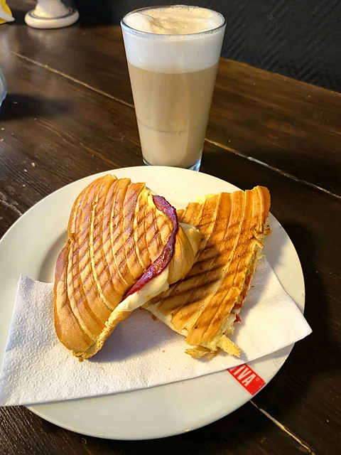 Getoastetes Brioche-Croissant mit Schinken und Käse auf weißem Teller, dahinter ein Glas Galão mit Milchschaum auf dunkl