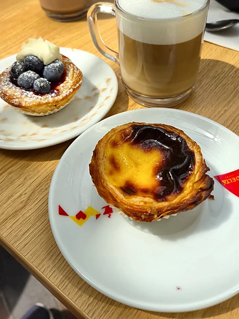 Pastel de Nata mit karamellisierter Oberfläche auf weißem Teller, daneben ein Cappuccino im Glas und ein Blaubeer-Törtch