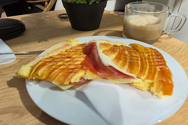 Getoastetes Croissant mit Schinken und Käse auf einem Teller, dahinter ein Glas Eiskaffee und ein Blumenstrauß auf dem H