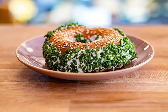 Bagel mit Frischkäse, rundum in Schnittlauch gewälzt, auf rosa Teller vor unscharfer Theke