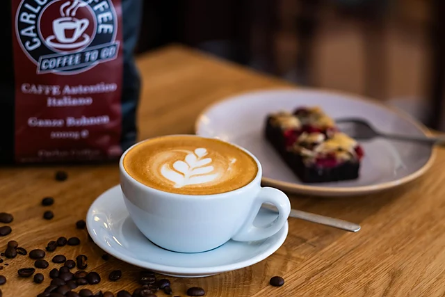 Cappuccino mit Latte-Art neben einem Stück Brownie mit Beeren, dahinter eine Packung Carlos Coffee Bohnen und verstreute Kaffeebohnen