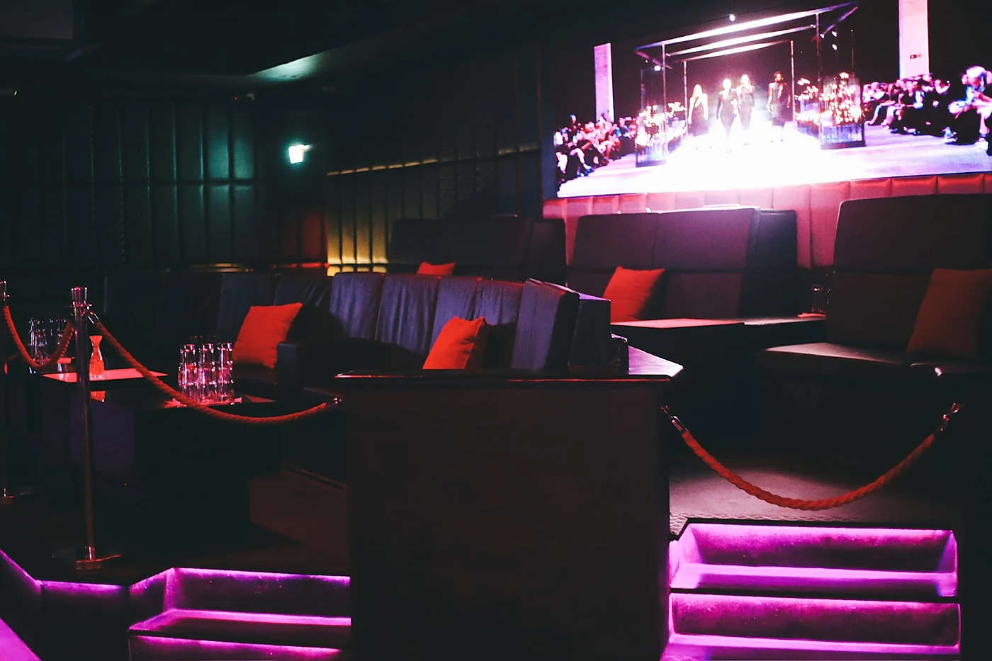 Lounge mit dunklen Sofas, roten Kissen, Absperrkordeln und violett beleuchteten Stufen