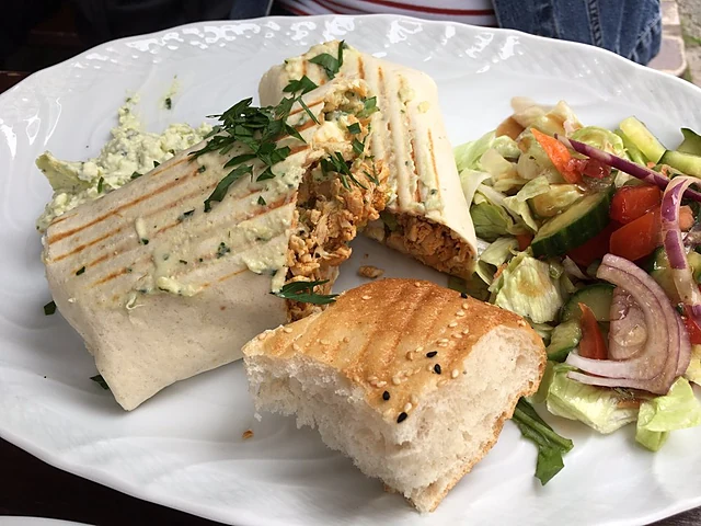 Gegrilltes Panini mit Hähnchen und Kräutern, dazu Salat mit Tomate, Gurke und roten Zwiebeln auf weißem Teller