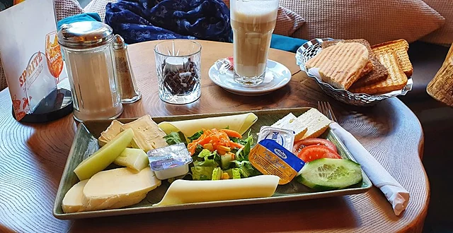 Frühstücksplatte mit Käse, Gemüse und Butter auf einem Holztisch, dahinter Latte macchiato und ein Korb mit Toast