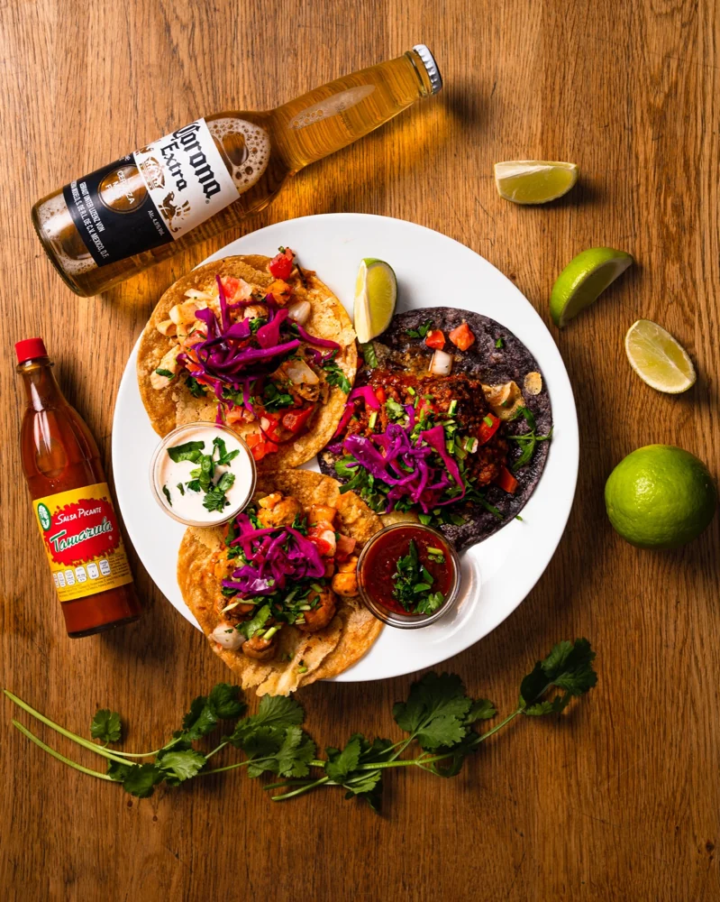 Drei Tacos mit Rotkohl, Koriander und Salsa auf weißem Teller, daneben Limetten, Hot Sauce und eine Flasche Corona
