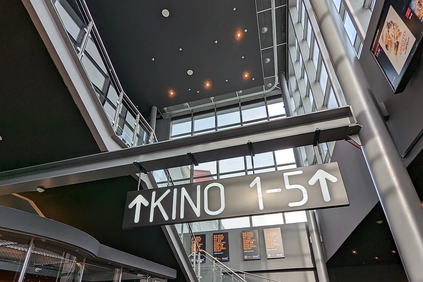 Hängendes Leuchtschild mit der Aufschrift Kino 1-5 und Pfeilen unter einer Stahl-Glas-Decke im Kinofoyer