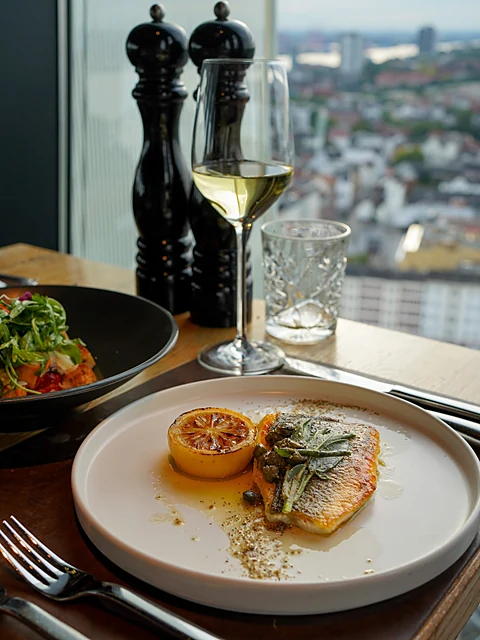 Fischgericht mit Zitrone auf weißem Teller, daneben ein Glas Weißwein und der Blick über Hamburg