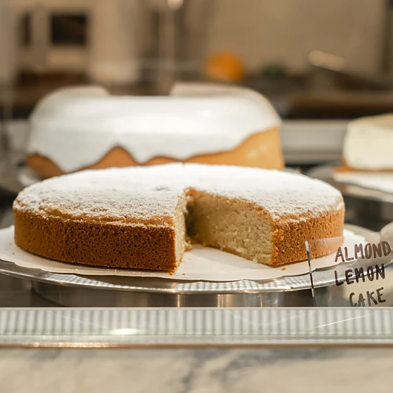 Angeschnittener Almond Lemon Cake mit Puderzucker in der Kuchenvitrine, dahinter ein glasierter Kuchen