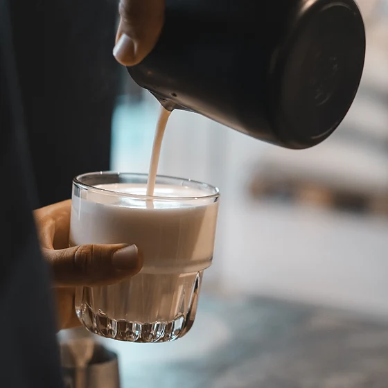 Aufgeschäumte Milch wird aus einem Kännchen in ein Glas mit Kaffee gegossen