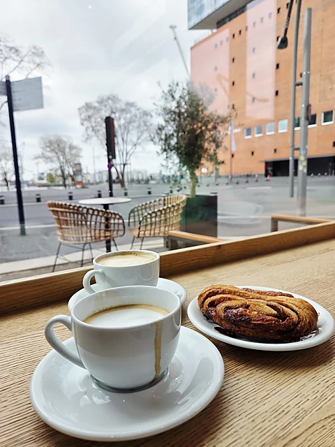 Espresso in weißer Tasse und ein Franzbrötchen auf einem Holztresen am Fenster, draußen Terrassenstühle