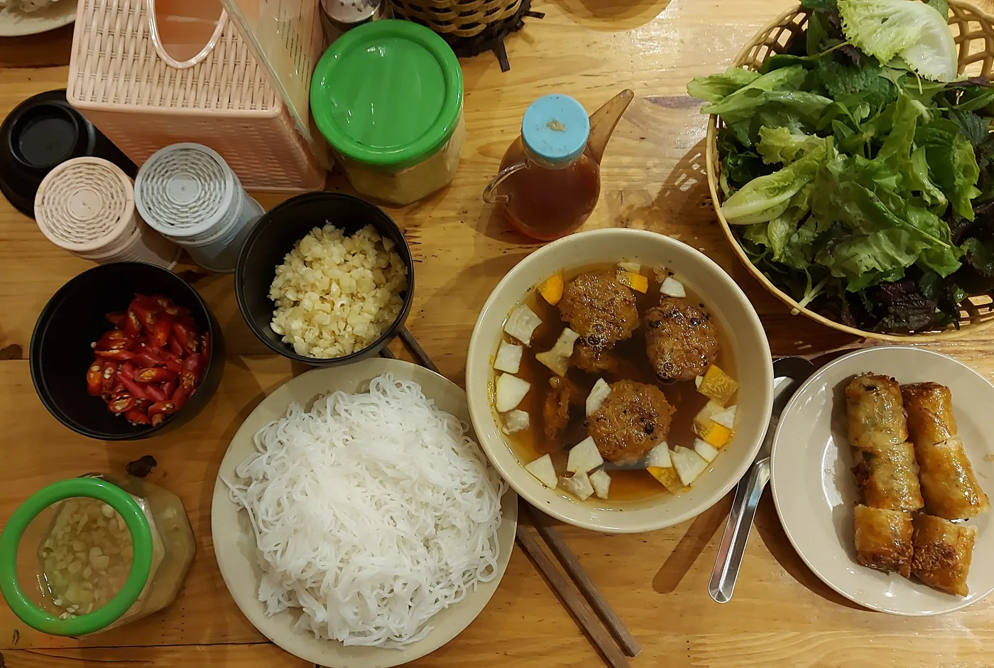 Bún chả mit Reisnudeln, gegrillten Fleischbällchen in Brühe, Frühlingsrollen und einem Korb frischer Kräuter