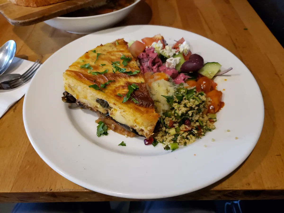 Stück Moussaka mit Béchamelhaube neben Salat auf einem weißen Teller