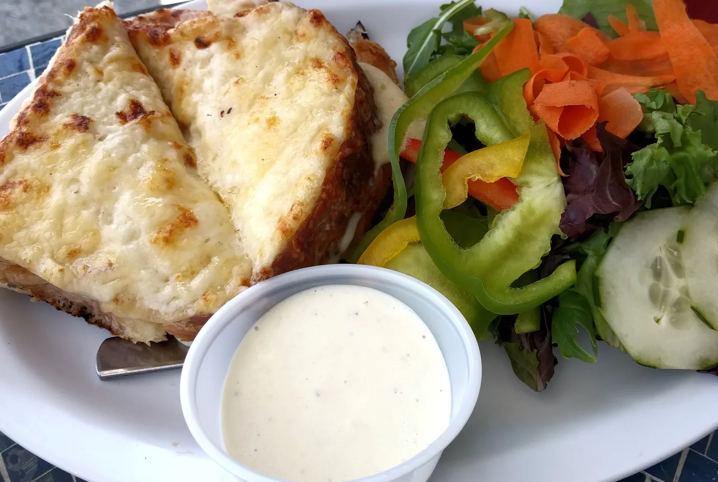 Aufgeschnittenes Croque mit geschmolzenem Käse, daneben ein Salat mit Paprika, Gurke und Karotte und ein Becher Dressing