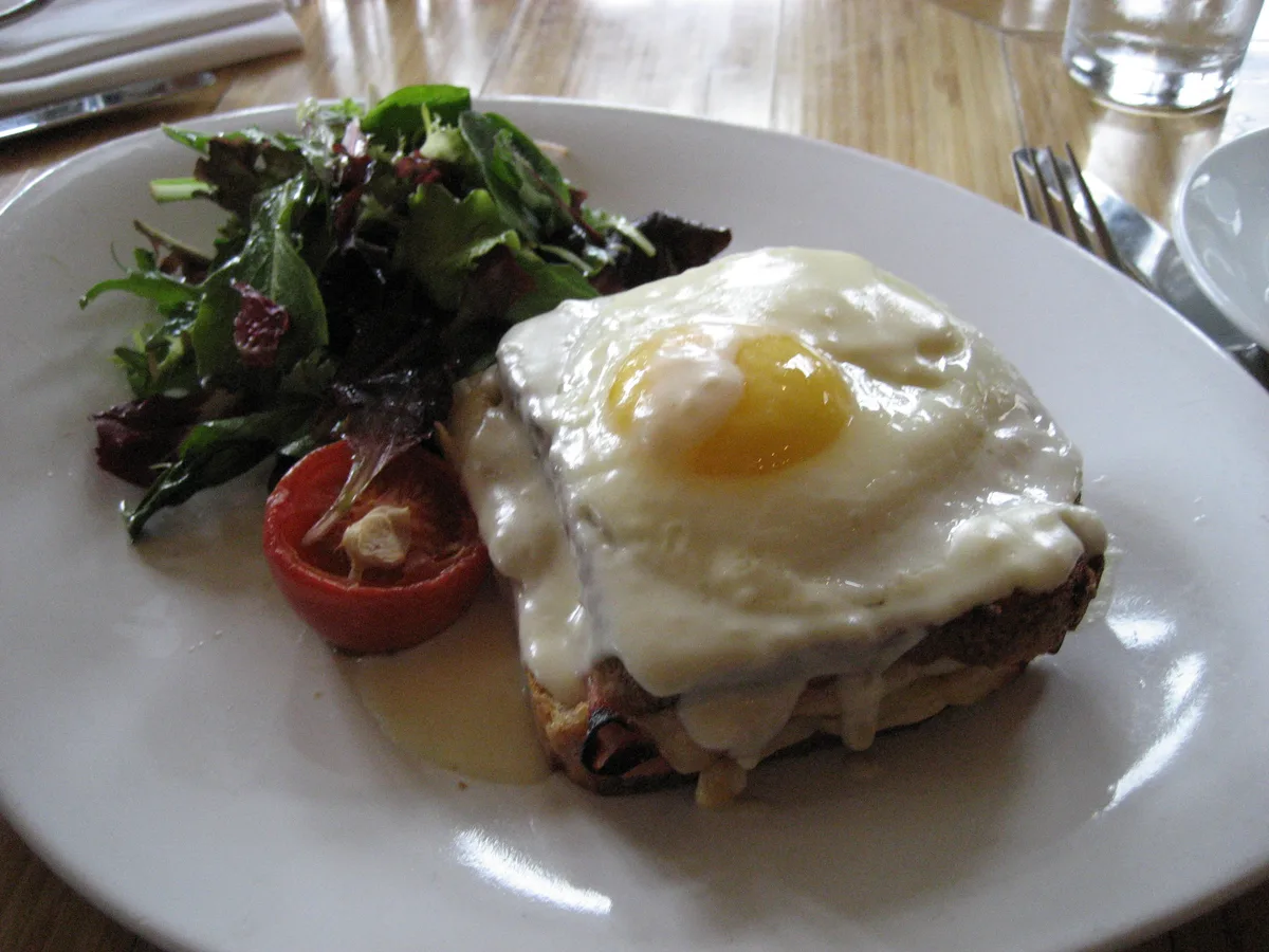 Croque Madame mit Spiegelei und geschmolzenem Käse, daneben Blattsalat und eine gebratene Tomate