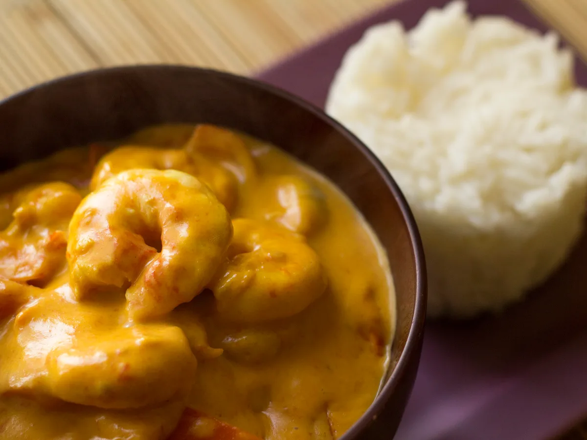 Gelbes Thaicurry mit Garnelen in einer dunklen Schale, daneben eine Portion Reis