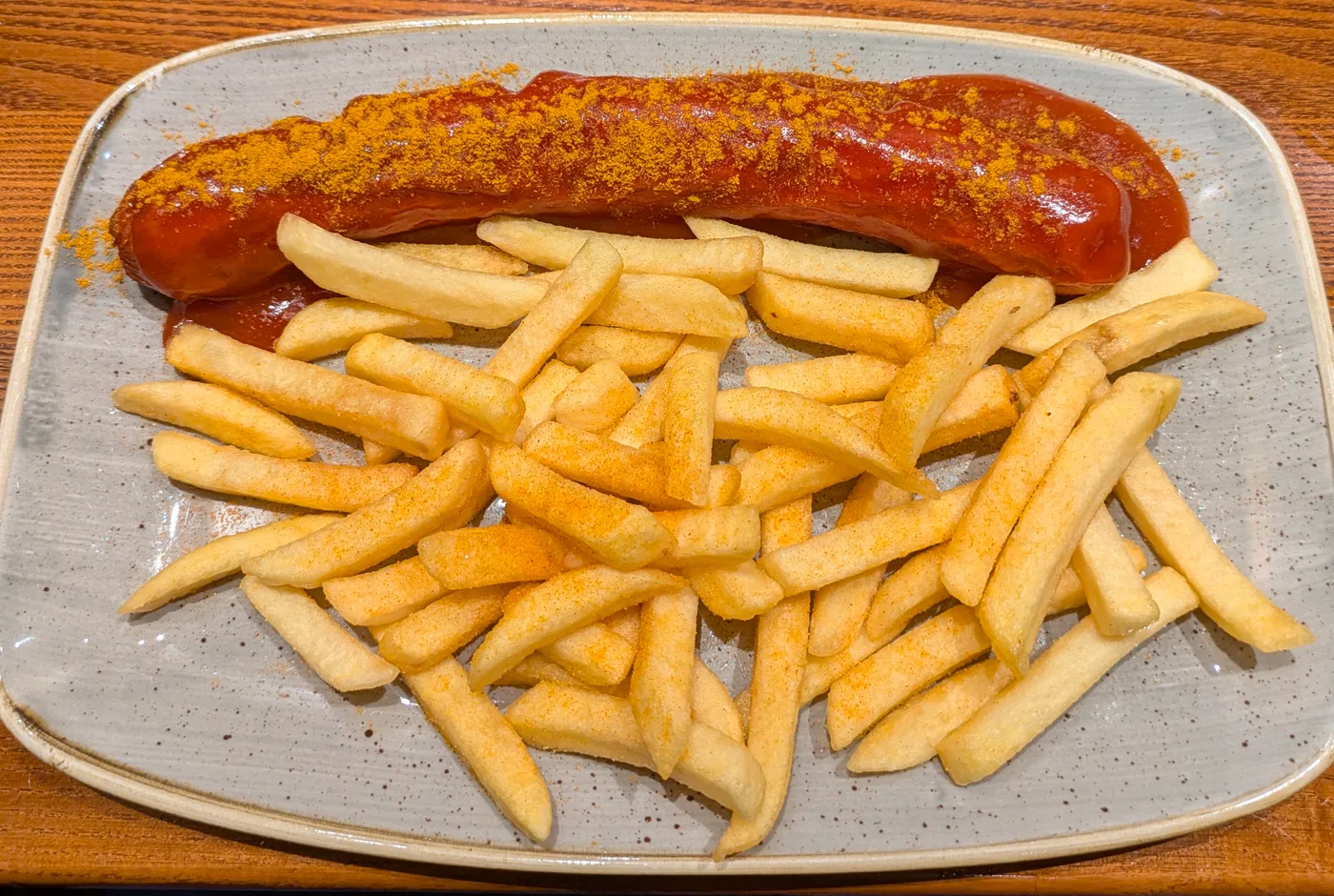 Currywurst mit Sauce und Currypulver, daneben Pommes auf einem hellen Teller