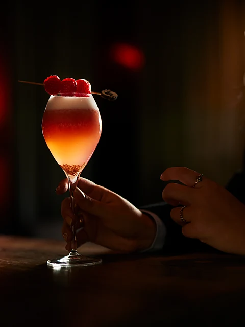 Rosafarbener Cocktail mit Kirschen im Stielglas, gehalten von einer Hand im gedämpften Licht der Bar