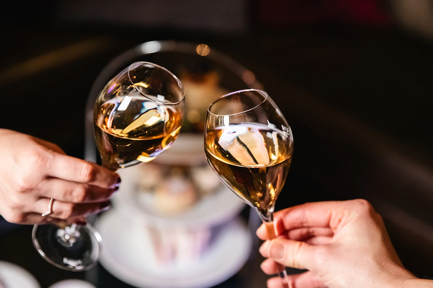 Zwei Hände stoßen mit Champagnergläsern an, im Hintergrund die Etagere des Afternoon Tea