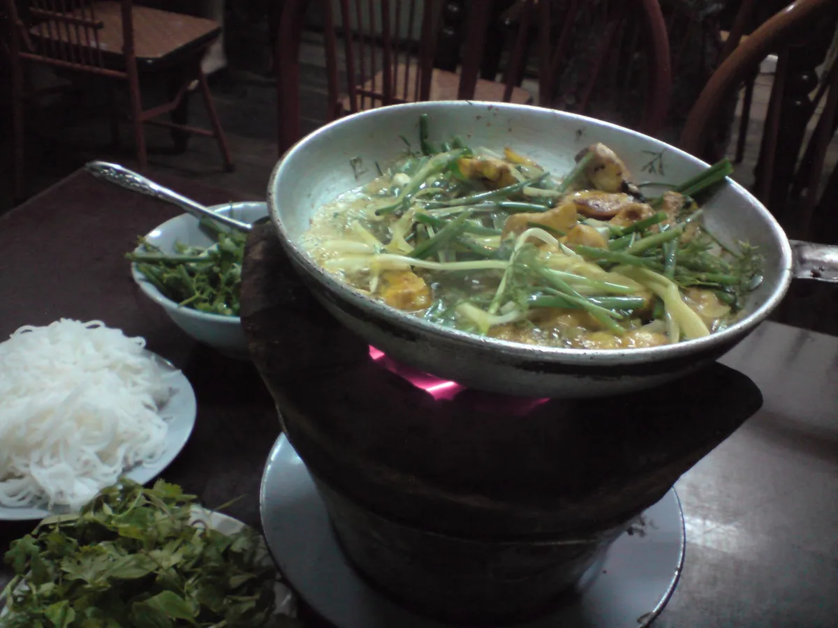 Vietnamesisches Fischgericht mit Kurkuma, Frühlingszwiebeln und Dill in einer Pfanne, daneben Reisnudeln und Kräuter