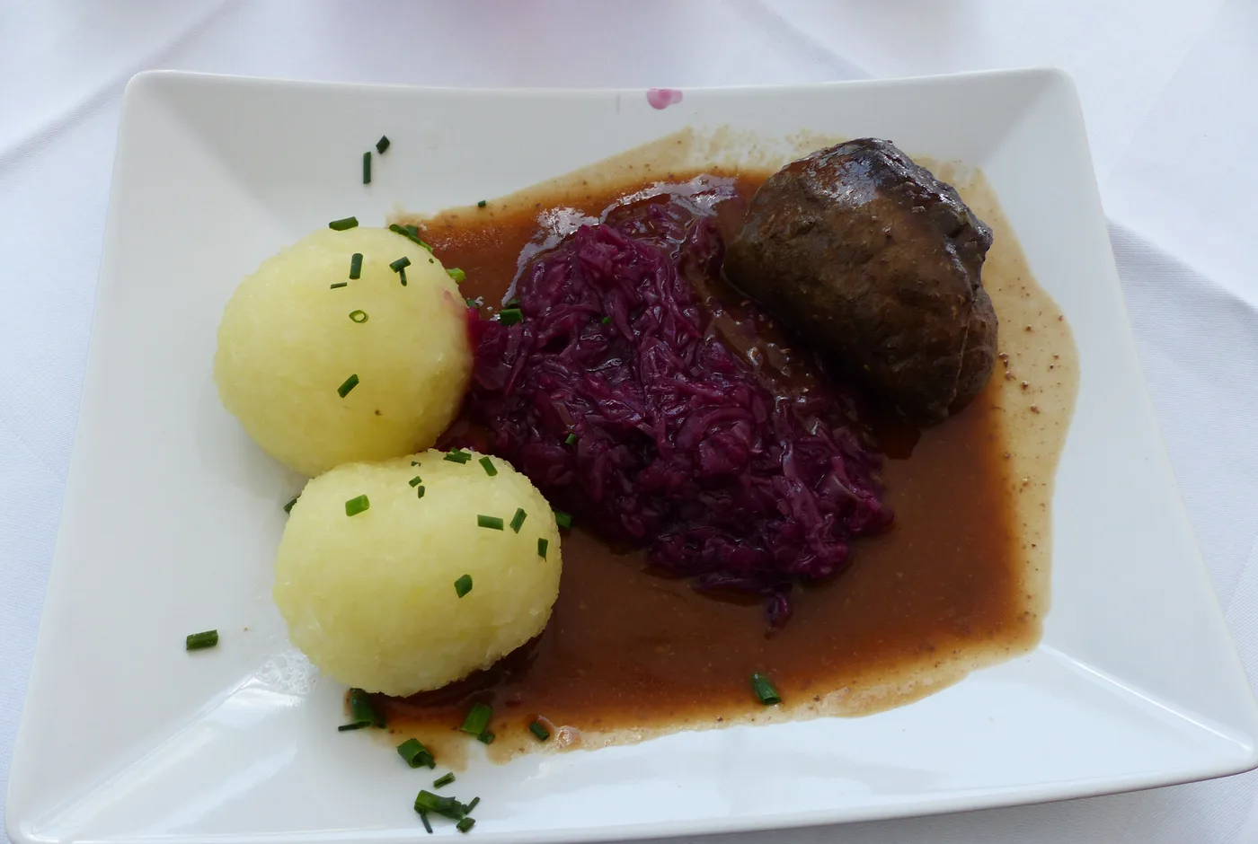 Rinderroulade mit Rotkohl, zwei Kartoffelklößen und dunkler Sauce auf einem weißen Teller