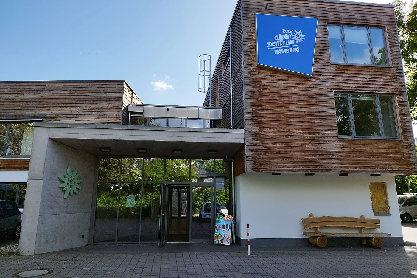 Eingang eines Holz- und Betongebäudes mit blauem Schild DAV alpinzentrum Hamburg, davor eine Holzbank