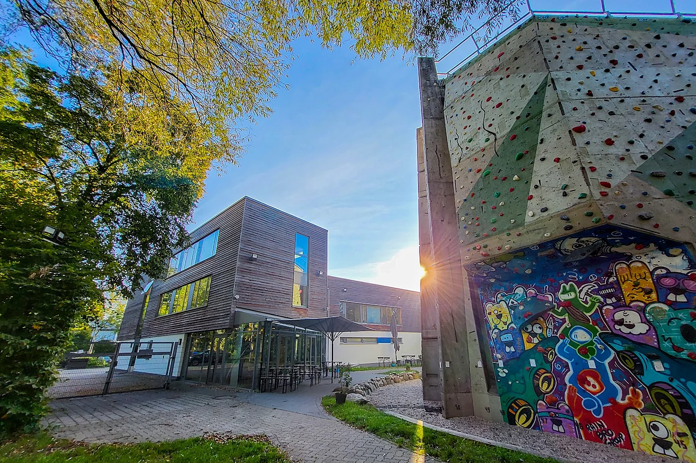Außengelände mit Holzgebäude, Terrasse und einer bunt mit Graffiti bemalten Betonwand mit Klettergriffen