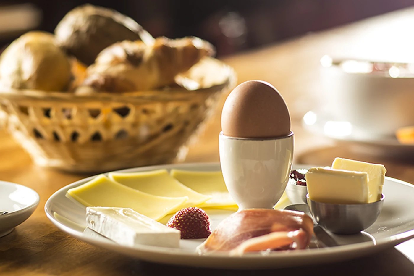 Frühstücksteller mit Ei im Becher, Käse, Schinken und Butter, dahinter ein Brotkorb