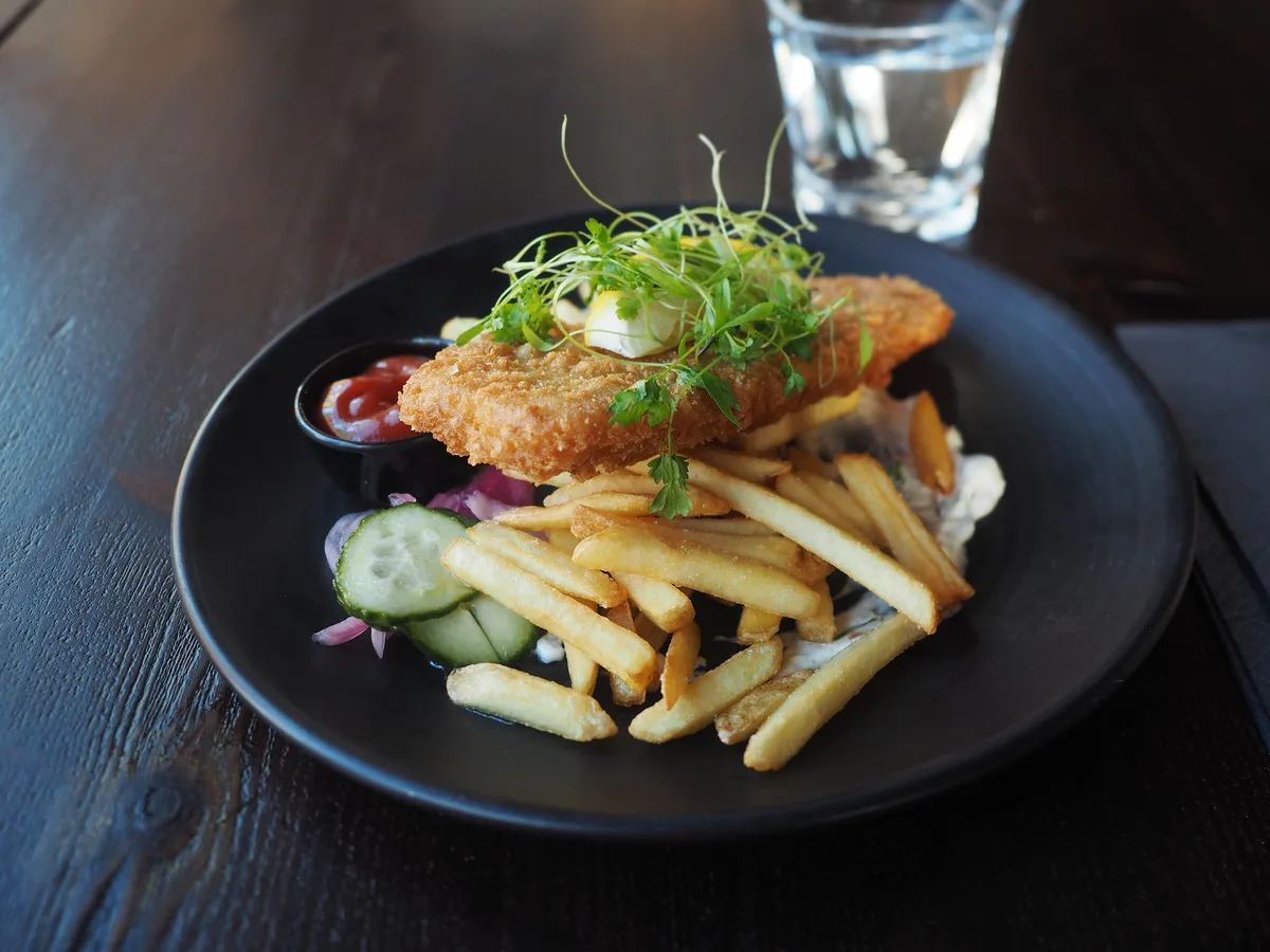 Paniertes Fischfilet mit Pommes frites, Gurke und Ketchup auf einem schwarzen Teller